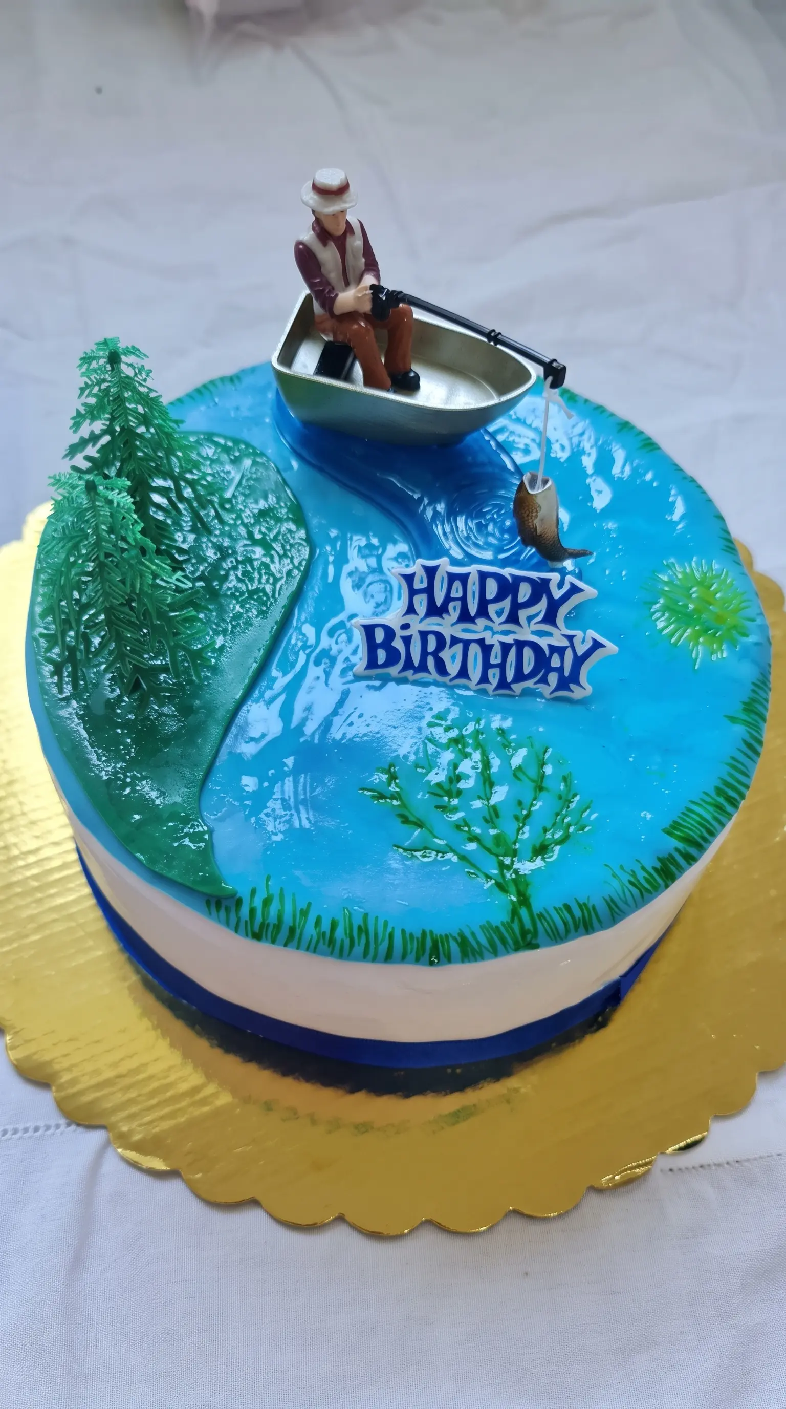 Birthday cake decorated with a fishing scene, boat topper, lake details and greenery